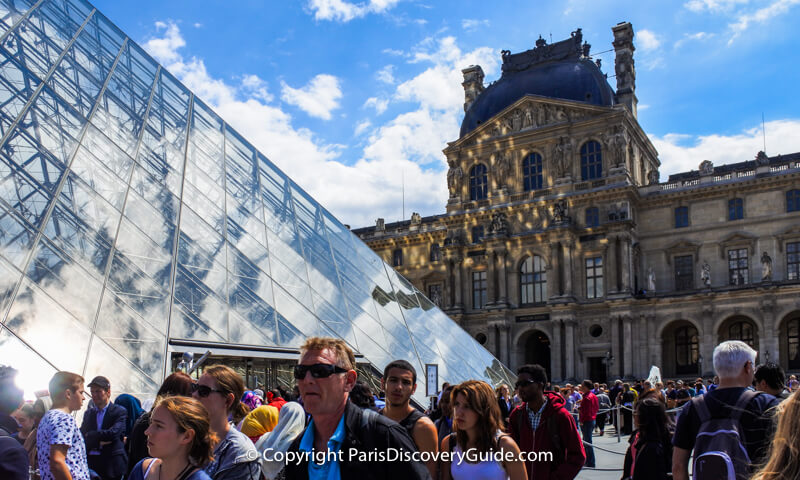 Ticket line at the Louvre winding around the courtyard; nearby signs warn of a 2+ hour wait 