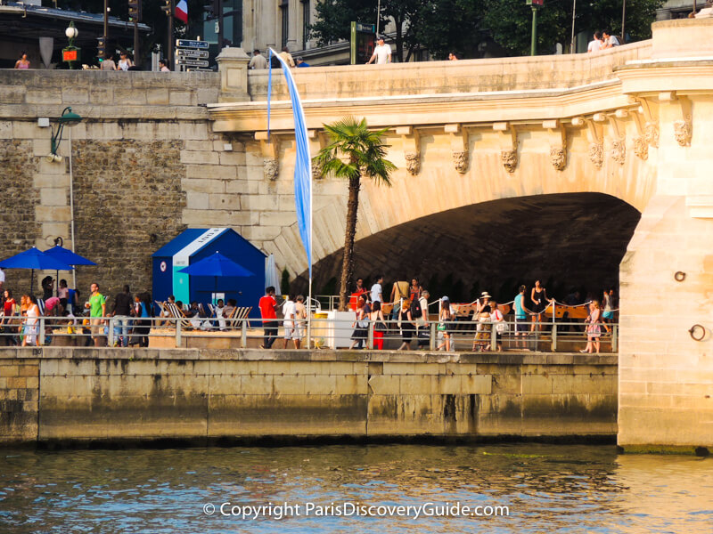 Palm tree, beach umbrellas, and striped beach chairs next to the Pont Neuf Bridge at Paris Plages 