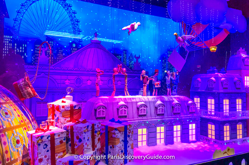 A pigeon and a dove embark on a magical journey as animated doll figures play musical instruments and dance on a Parisian rooftop with a giant ferris wheel and a hot air balloon in the background - part of a multi-window scene at Galeries Lafayette A pigeon and a dove embark on a magical journey as animated doll figures play musical instruments and dance on a Parisian rooftop with a giant ferris wheel and a hot air balloon in the background - part of a multi-window scene at Galeries Lafayette