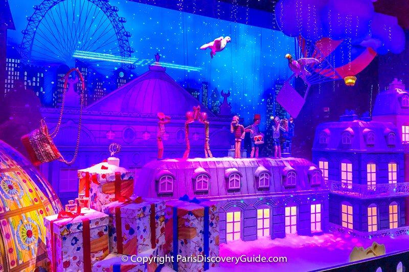 A pigeon and a dove embark on a magical journey as animated doll figures play musical instruments and dance on a Parisian rooftop with a giant ferris wheel and a hot air balloon in the background - part of a multi-window scene at Galeries Lafayette A pigeon and a dove embark on a magical journey as animated doll figures play musical instruments and dance on a Parisian rooftop with a giant ferris wheel and a hot air balloon in the background - part of a multi-window scene at Galeries Lafayette